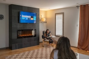 Un salon avec un téléviseur à écran plat fixé sur un mur sombre, une cheminée en dessous et un sol à carreaux.