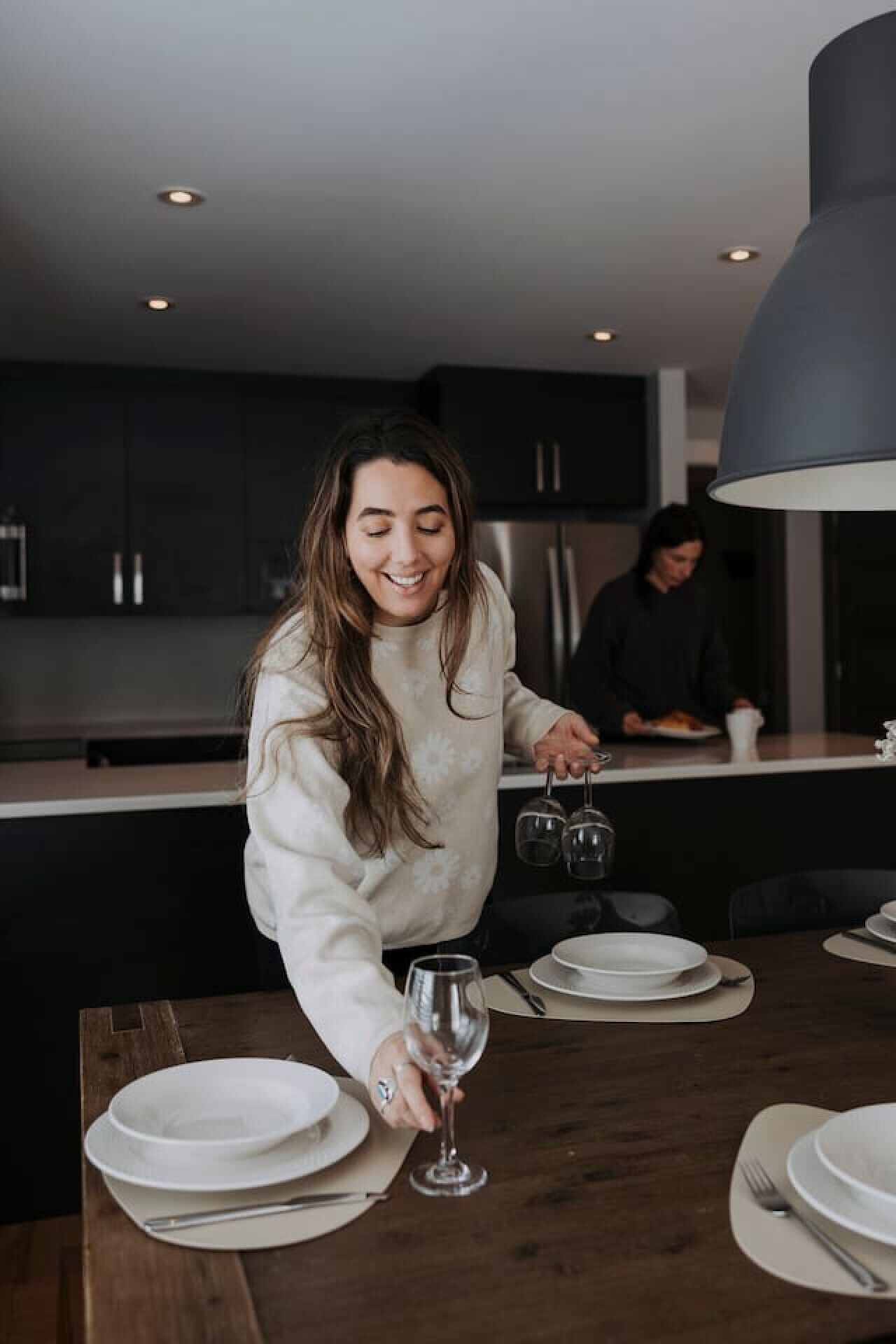 Una persona con suéter blanco ordenando copas de vino sobre una mesa de madera en una cocina moderna con alacenas oscuras y una lámpara colgante grande.