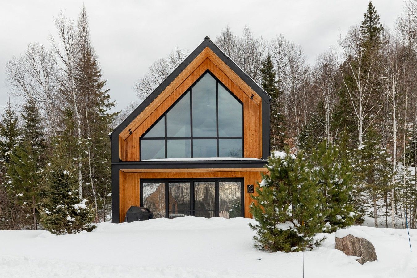 A modern two-story cabin with large windows and a wooden exterior, surrounded by snow-covered trees.
