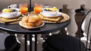 Una mesa redonda con alimentos para el desayuno, incluyendo cruasanes, fruta y jugo de naranja, en un balcón con sillas y barandilla.