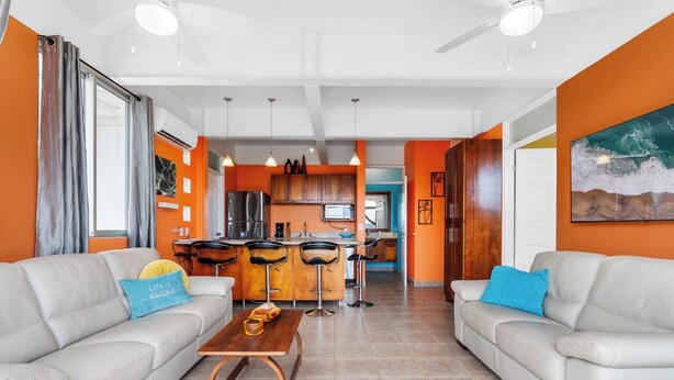 A modern living room with two sofas, a coffee table, and a kitchen in the background. - Modern Top-Floor Condo with Panoramic Views and Community Pool (Sardinal)