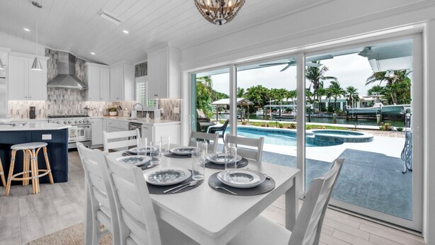 A modern kitchen with white cabinetry, a large island, and a dining area with a view of a pool and tropical landscape.