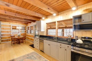 A log cabin interior with a wooden dining table, chairs, a kitchen with a stove, sink, and cabinets.