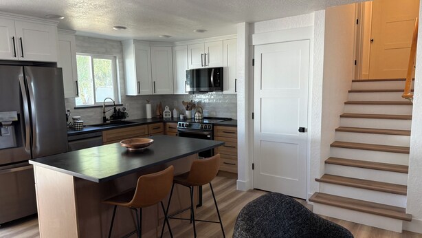 A modern kitchen with white cabinets, a stainless steel refrigerator, a microwave, and a stove. There are two bar stools and a round table. A wooden staircase is visible to the right. - Modern Upscale Hermiston Home • Pet-Friendly • Private Fenced Yard (Hermiston)