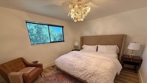 A bedroom with a large bed, a nightstand, a chair, a patterned rug, and a window with a view of trees. - Modern Upscale Hermiston Home • Pet-Friendly • Private Fenced Yard (Hermiston)