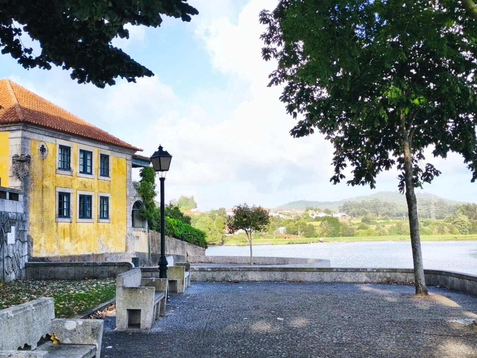 A historic building with a yellow facade and a red-tiled roof, situated next to a body of water with a view of hills in the distance.