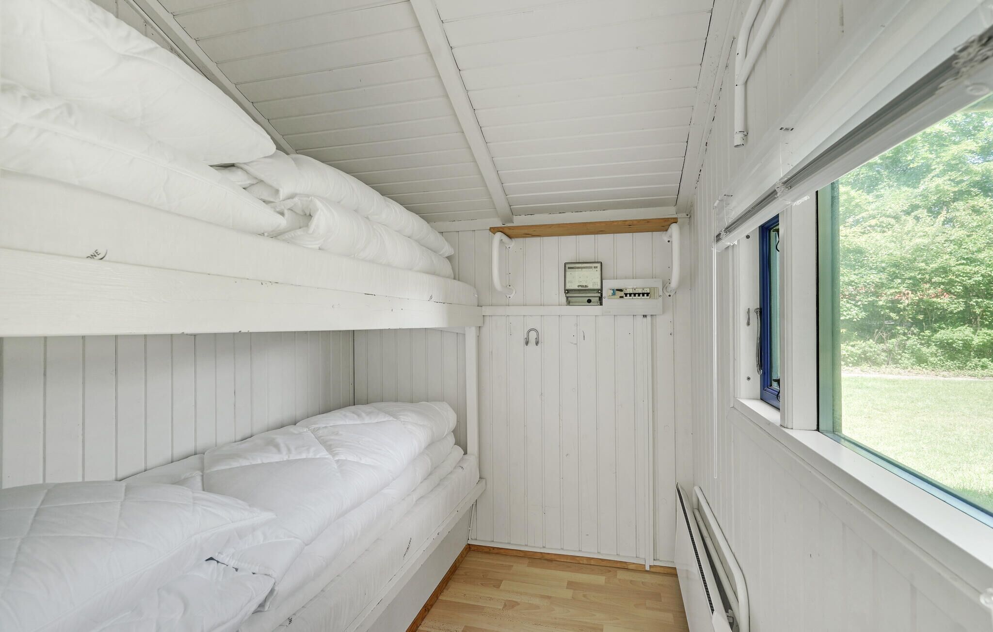 Une petite pièce de style cabane, aux murs blancs, avec des lits superposés, une petite étagère sur laquelle se trouve une radio et une fenêtre donnant sur la verdure.
