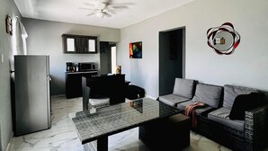 A living room with a sofa, a coffee table, a dining table, a kitchen area, and a ceiling fan.