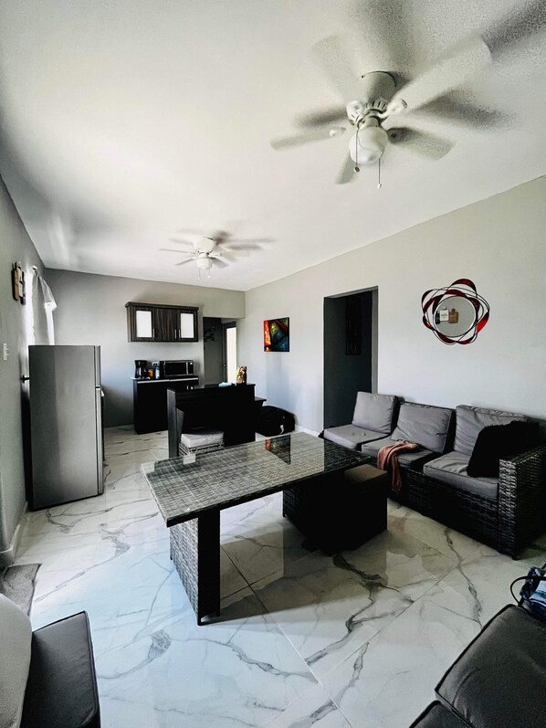 A living room with a sofa, a coffee table, a dining table, a kitchen area, and a ceiling fan.