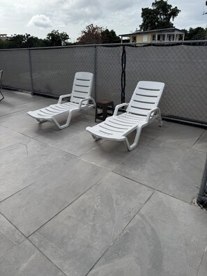 Two white lounge chairs on a concrete patio with a chain-link fence in the background.