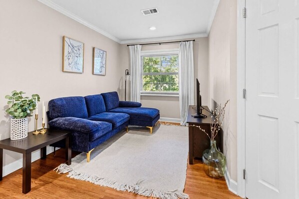 Un salon avec un canapé bleu, un tapis blanc, une table d’appoint avec une plante et une fenêtre avec des rideaux.