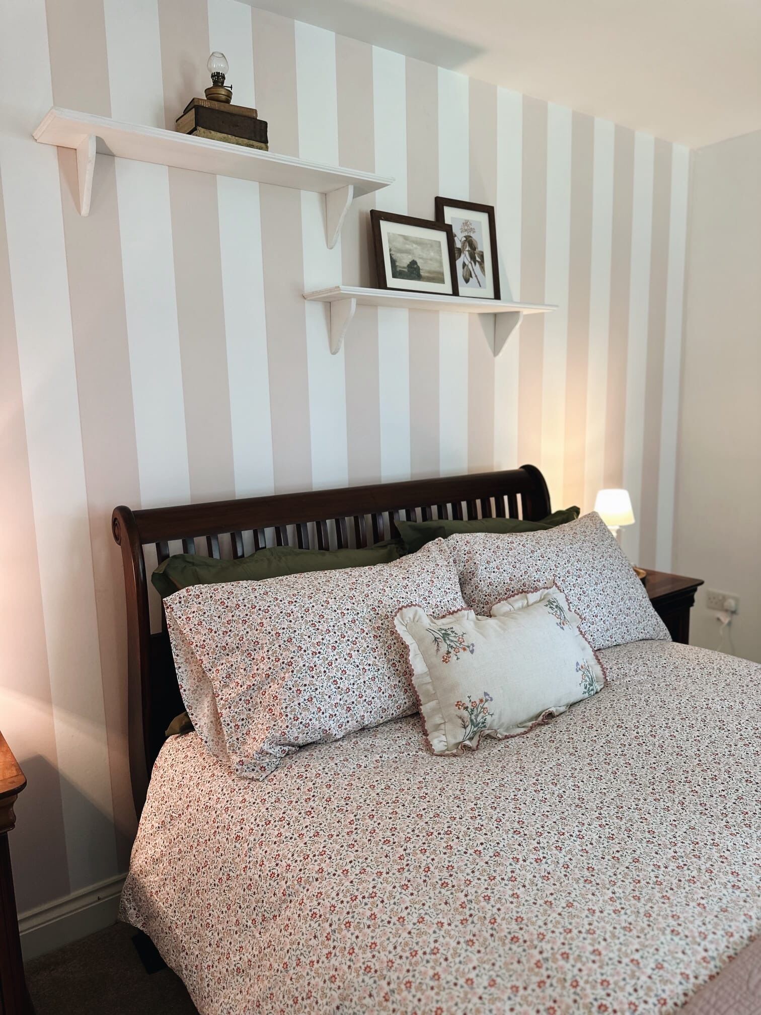 A bedroom with a striped wall, a wooden bed frame, floral bedding, and two wall-mounted shelves with pictures.
