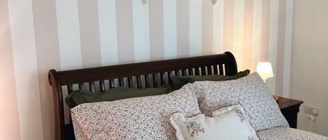 A bedroom with a striped wall, a wooden bed frame, floral bedding, and two wall-mounted shelves with pictures.
