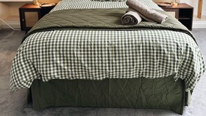 Cama bem arrumada com colcha xadrez e almofadas, duas mesas de cabeceira com abajures, e uma estante com livros e cestos.