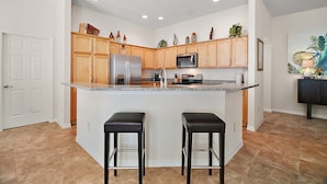 A modern kitchen with wooden cabinets, a stainless steel refrigerator, a microwave, and a granite countertop. There are two black bar stools and a small island with a sink. - Charming 3-bedroom villa walking distance to Lake Sumter Sq  Golf Cart included (The Villages)