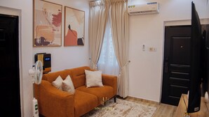 A living room with a sofa, a fan, a TV, and a window with curtains. - Akoka Oasis 1-Bed Near Unilag (Lagos)