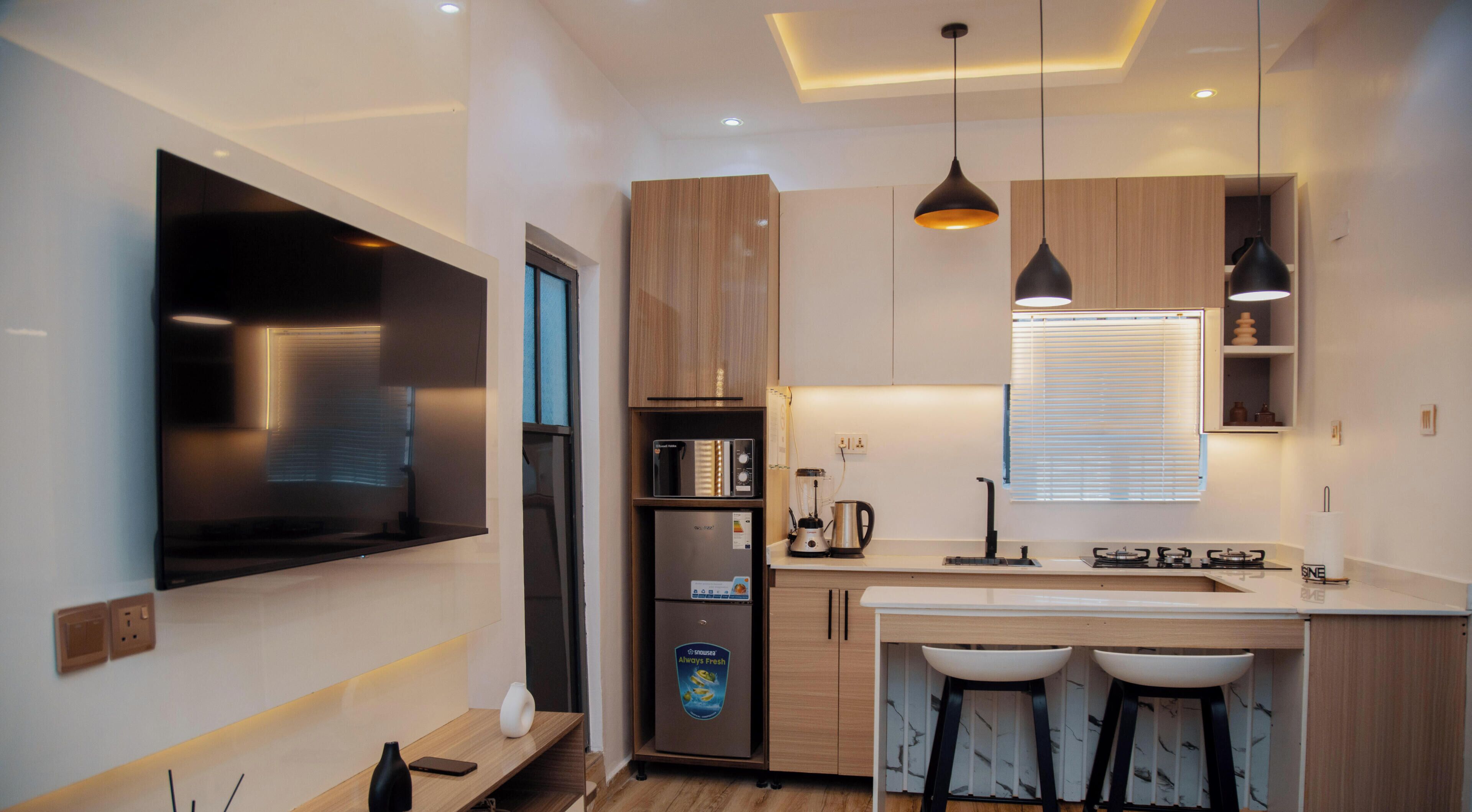 A modern kitchen with a built-in TV, wooden cabinets, a refrigerator, and a bar area with stools. - Akoka Oasis 1-Bed Near Unilag (Lagos)