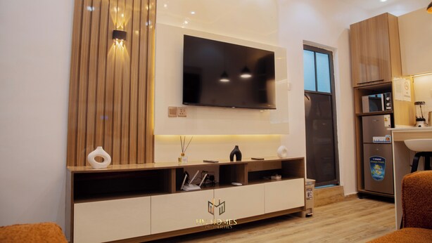 A modern living room with a flat-screen TV mounted on the wall, wooden paneling, a built-in entertainment unit, and a kitchen area with a refrigerator and sink. - Akoka Oasis 1-Bed Near Unilag (Lagos)