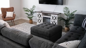 A modern living room with a large grey sectional sofa, a brown armchair, a flat-screen TV mounted on the wall, a ceiling fan, and two potted plants. - Meridian Sunset views w/playroom and private HOT TUB (Meridian)