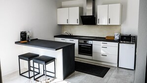 A modern kitchen with white cabinets, a black countertop, a built-in oven, a microwave, and a coffee maker. There are two black stools at the island. - New modern apartment, 74sqm, balcony, king-size box-spring bed (Offenburg)