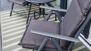 A round table with four chairs, each with a dark cushion and a metal frame. There are decorative items on the table, including a small vase and a plant with yellow flowers. The setting is on a balcony with a striped floor mat and a glass door leading to a building with a beige wall and a window with sheer curtains. - New modern apartment, 74sqm, balcony, king-size box-spring bed (Offenburg)