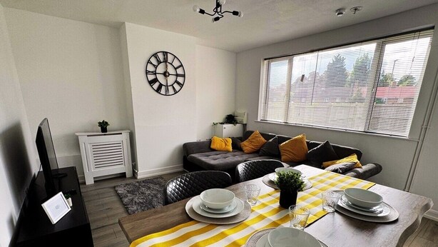 A modern living room with a dining area. There is a sofa with cushions, a dining table set with plates and glasses, and a clock on the wall. - Home In Northampton | Contractors Welcome (Northampton)