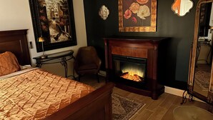 A bedroom with a bed, a fireplace, a chair, a table, and artwork on the walls.