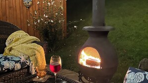 An outdoor patio with a wood-burning stove, string lights, a wicker table, and a cushioned bench.