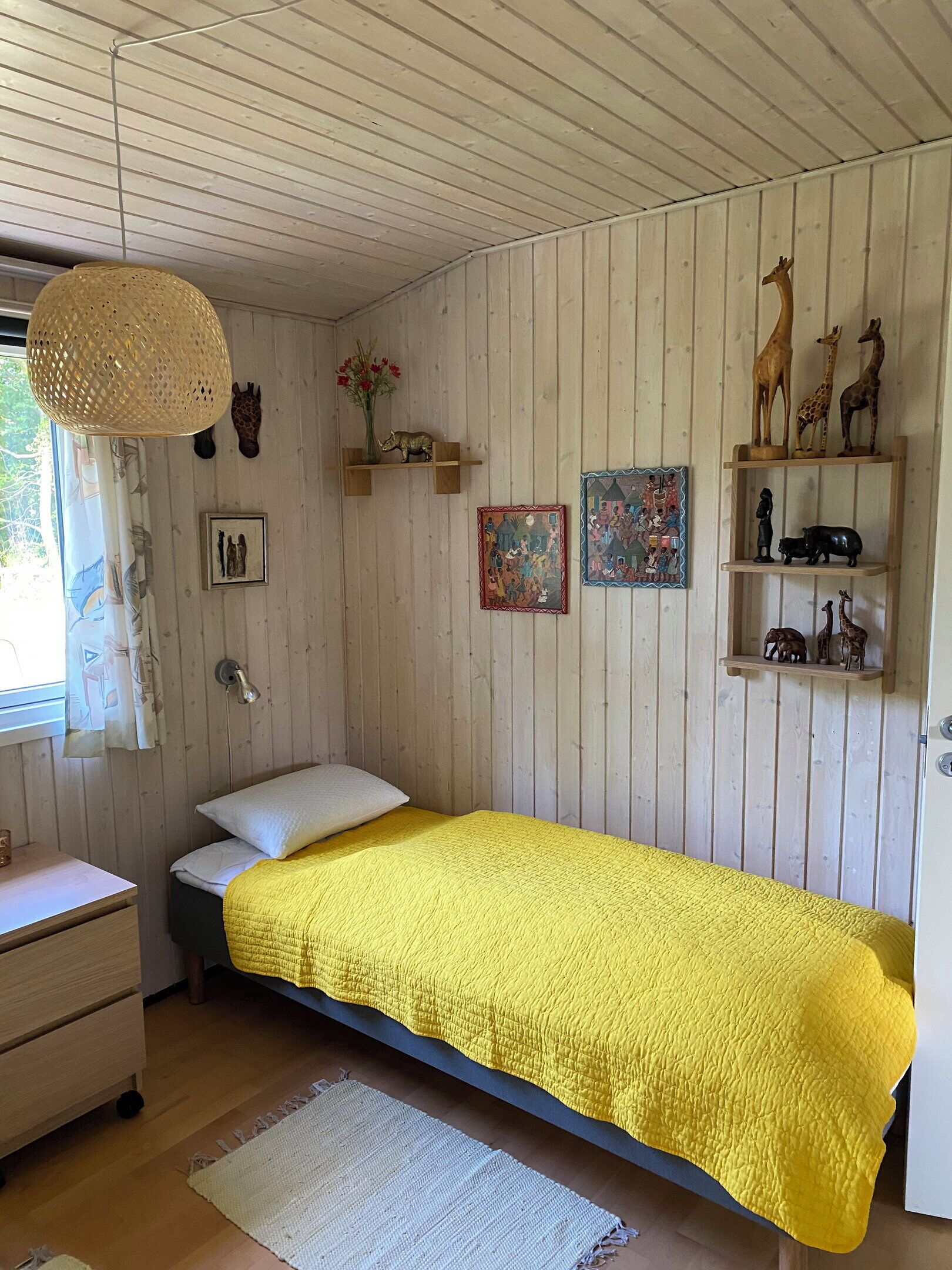 Een kamer met een bed, een bureau, een raam met gordijnen, een hanglamp en wanddecoraties, waaronder beeldjes van dieren en ingelijste foto's.