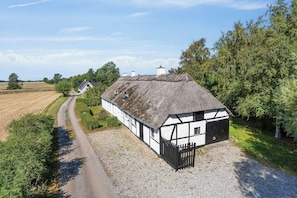 A traditional thatched-roof house with a gravel driveway, surrounded by fields and trees.