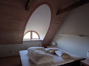 A bedroom with a sloped ceiling, a window, a bed with pillows, a bedside table, and a lamp.