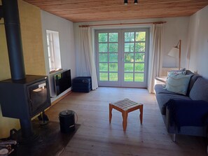 A room with a wood-burning stove, a sofa, a small table, and a large window with curtains.