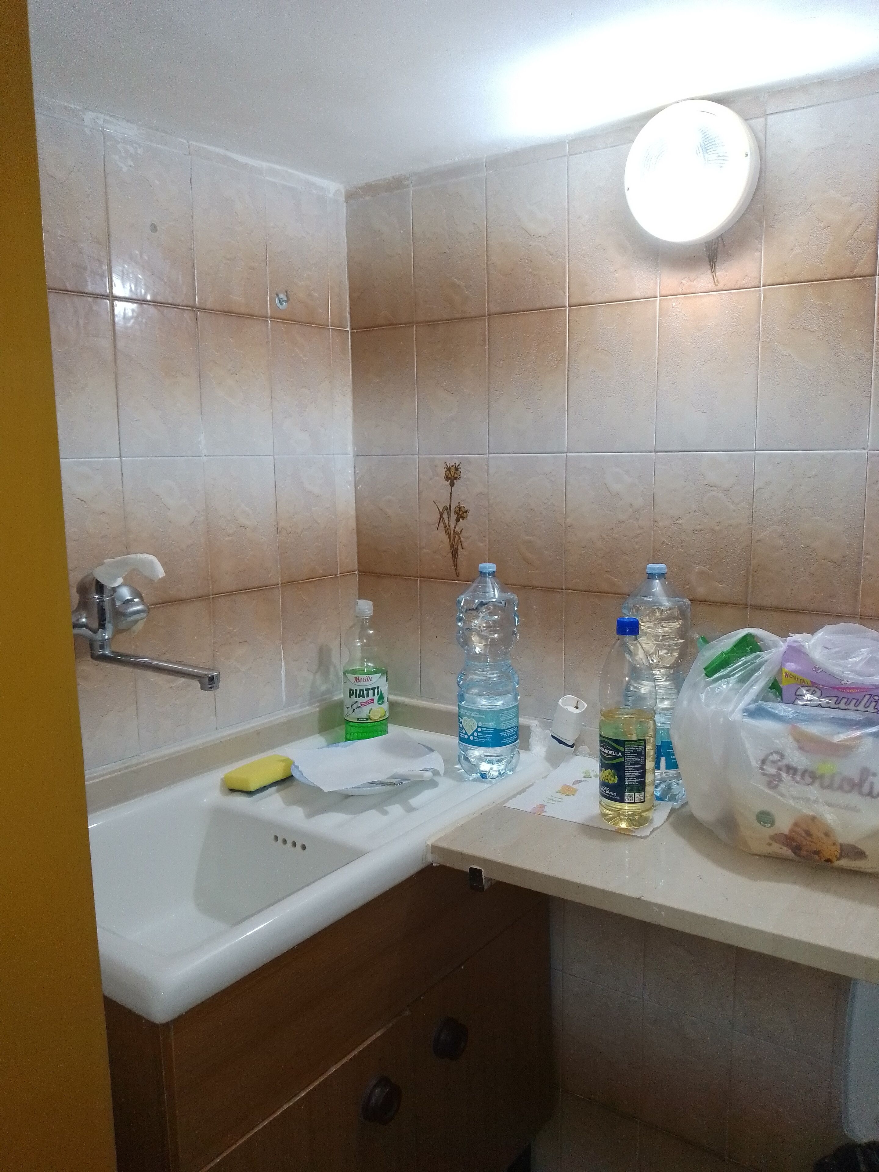 Een badkamer met een wastafel, een spiegel en een toilet. Er staan toiletartikelen op het aanrecht, waaronder een fles handzeep, een fles water en een zakje brood.