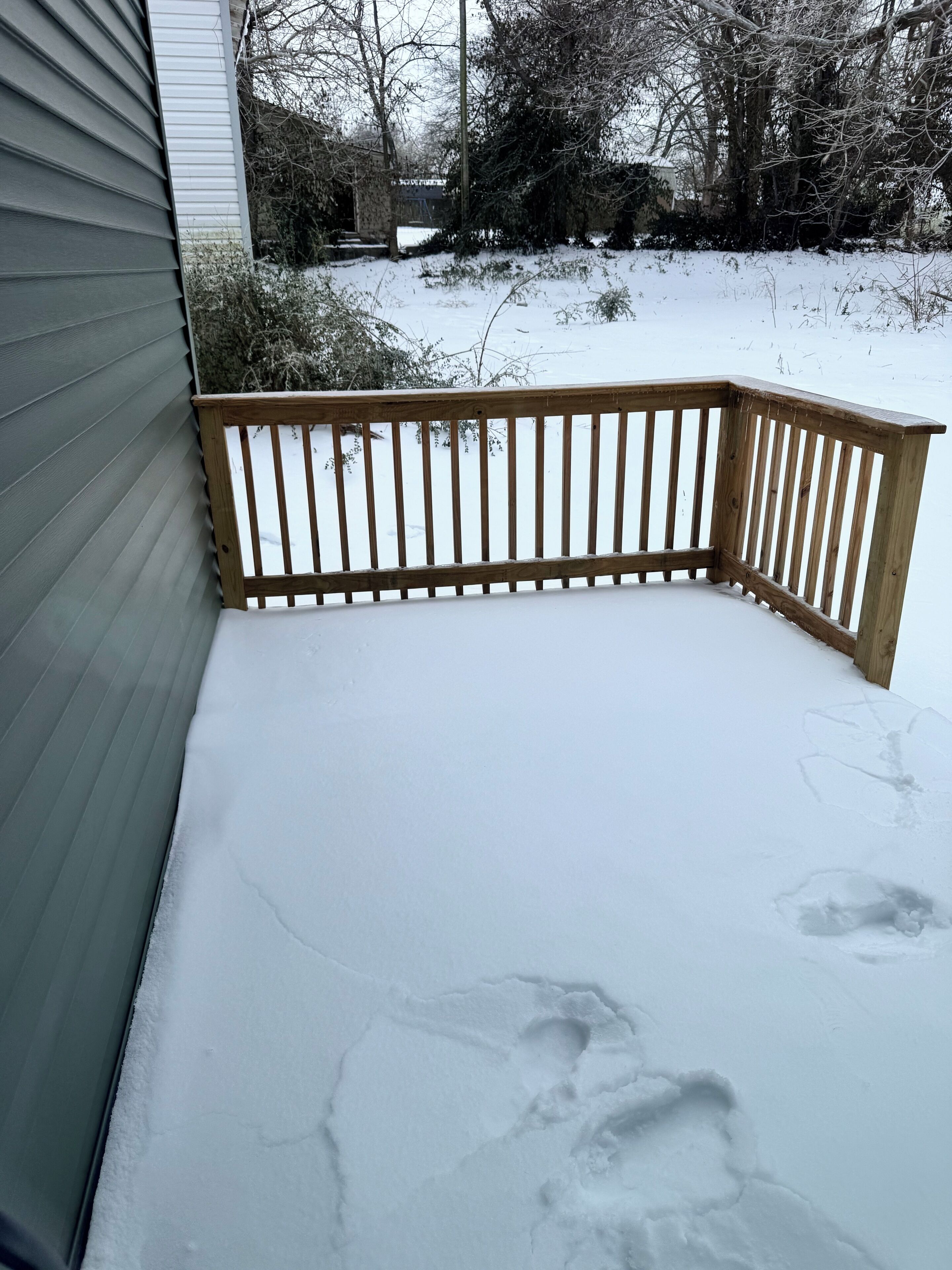 A snow-covered deck with a wooden railing, adjacent to a house with siding.