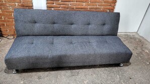 A grey upholstered sofa with a tufted backrest and four legs, placed on a concrete floor against a brick wall. - Comfortable and cozy apartment (Oaxaca de Juárez)