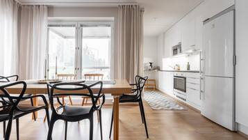 Une cuisine moderne avec des appareils électroménagers blancs, un coin repas avec une table et des chaises en bois, et de grandes fenêtres avec des rideaux.