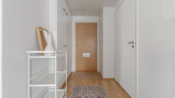 Un couloir étroit, avec une porte en bois au fond, une étagère blanche agrémentée d’un miroir et d’un cadre photo, et un tapis à motifs sur le sol.