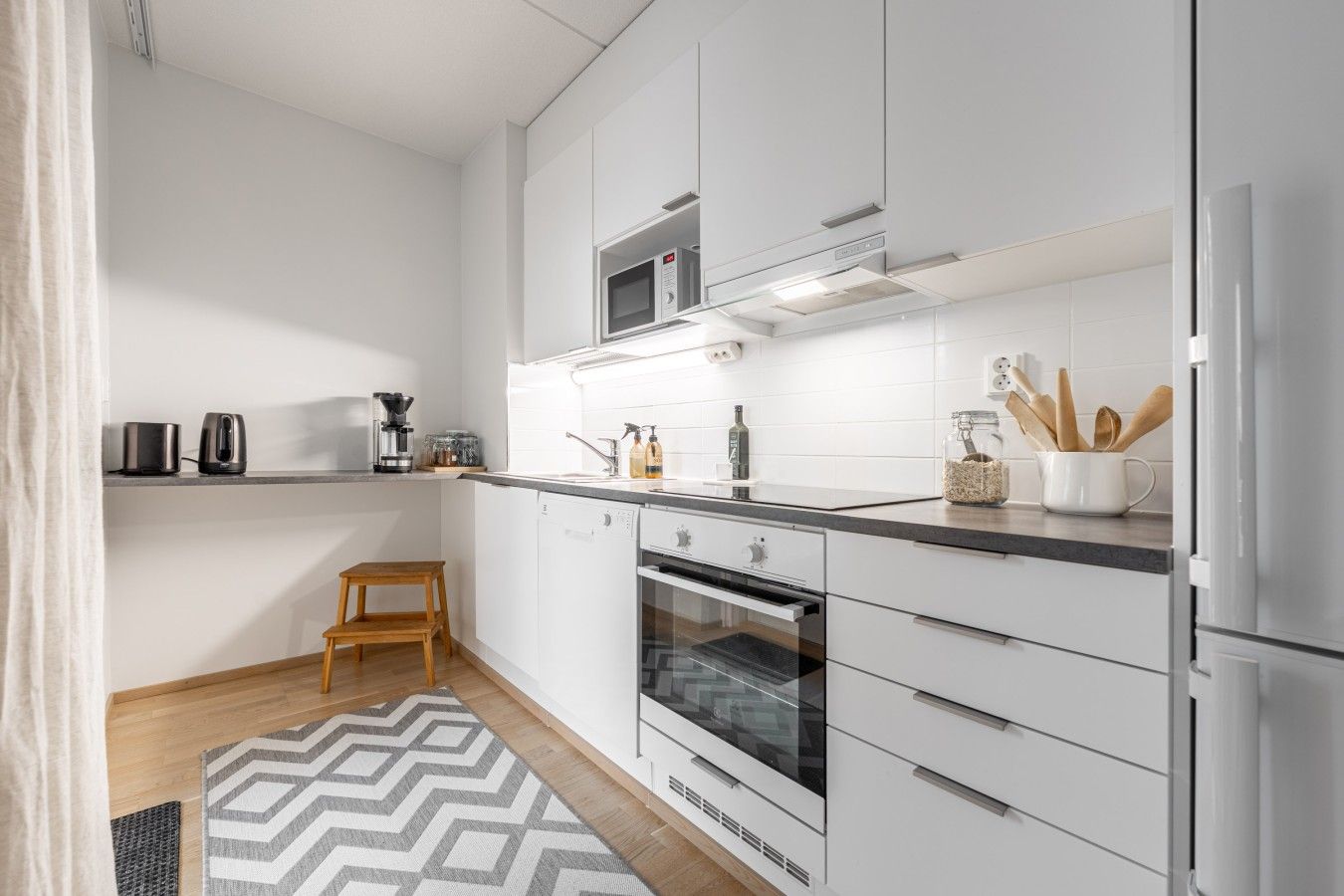 A modern kitchen with white cabinetry, a microwave, a coffee maker, and a small wooden stool.
