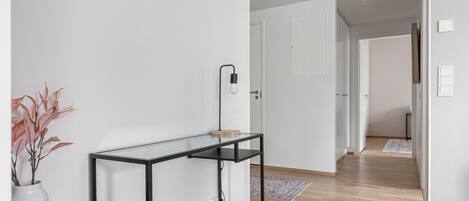 A hallway with a glass table, a white vase with dried plants, and a door leading to another room.