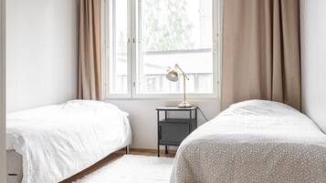 A bedroom with two beds, a window with curtains, a bedside table with a lamp, and a hanging light fixture.