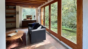 Un vaste espace de vie doté d’un plafond en bois, de grandes fenêtres donnant sur un jardin luxuriant, d’un canapé et d’une petite table sur laquelle repose un livre.