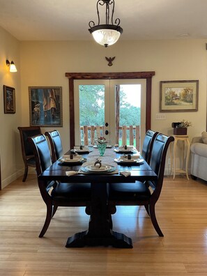 A dining room with a wooden table set for six, flanked by six chairs. There is a chandelier above the table, and the room has a view through a glass door to the outside.