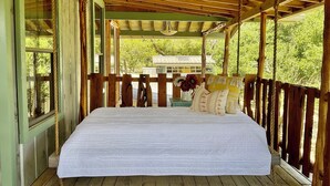 A wooden deck with a bed, a view of trees, and a house in the background.