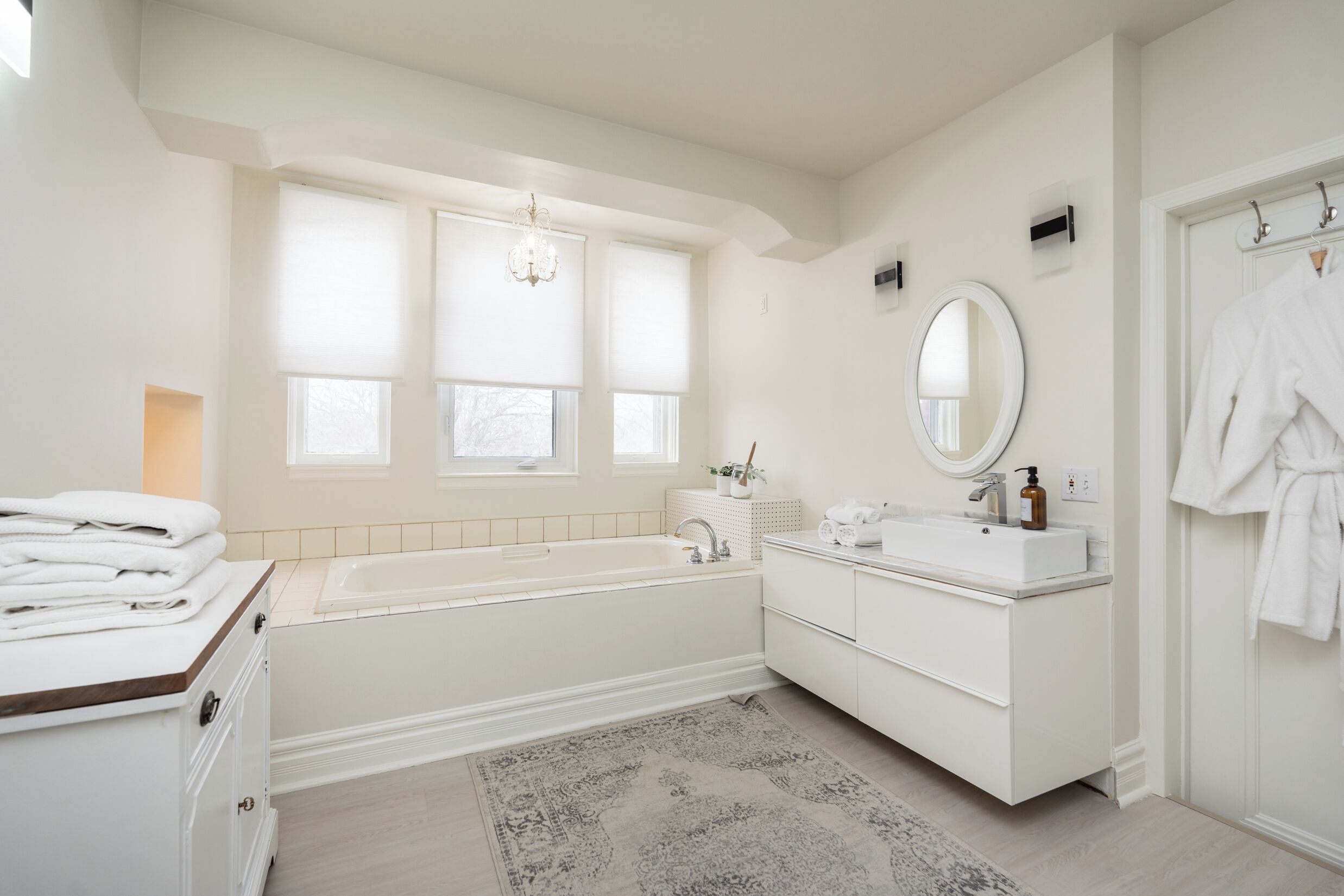 Une salle de bain moderne avec une baignoire, un lavabo et un miroir. Des serviettes sont pliées sur un meuble, et un peignoir est accroché à une porte.