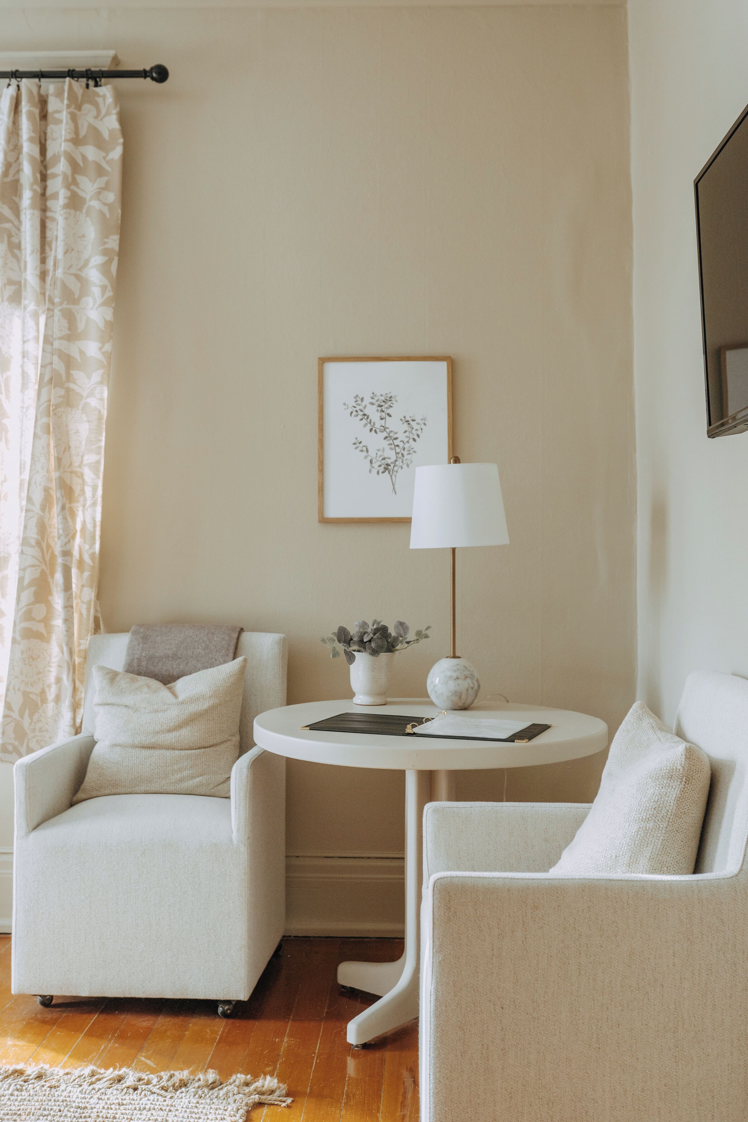Une pièce avec un mur de couleur beige, une œuvre d’art florale encadrée, une table ronde avec une lampe et deux chaises.