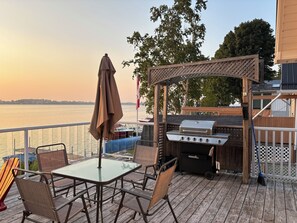 A deck with a glass table and chairs, a barbecue grill, and a view of the water.