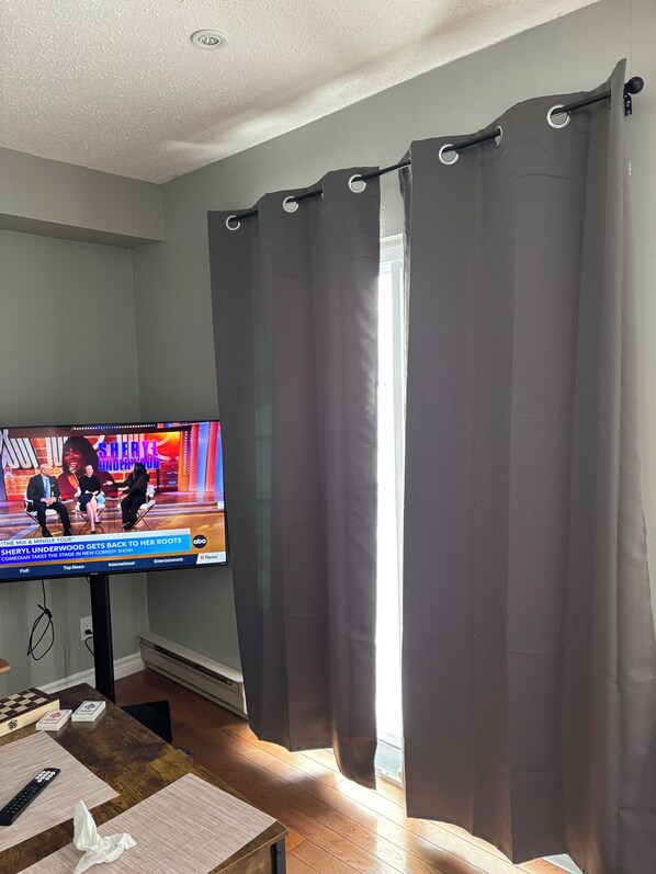 A living room with a television displaying a talk show, a wooden coffee table, and a window with curtains.