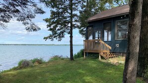 Una cabaña junto a un lago, con una terraza de madera y un amplio ventanal con vistas al agua.