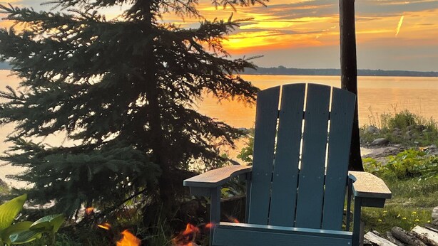 Una fogata con leña, una silla Adirondack azul y un atardecer sobre un lago.