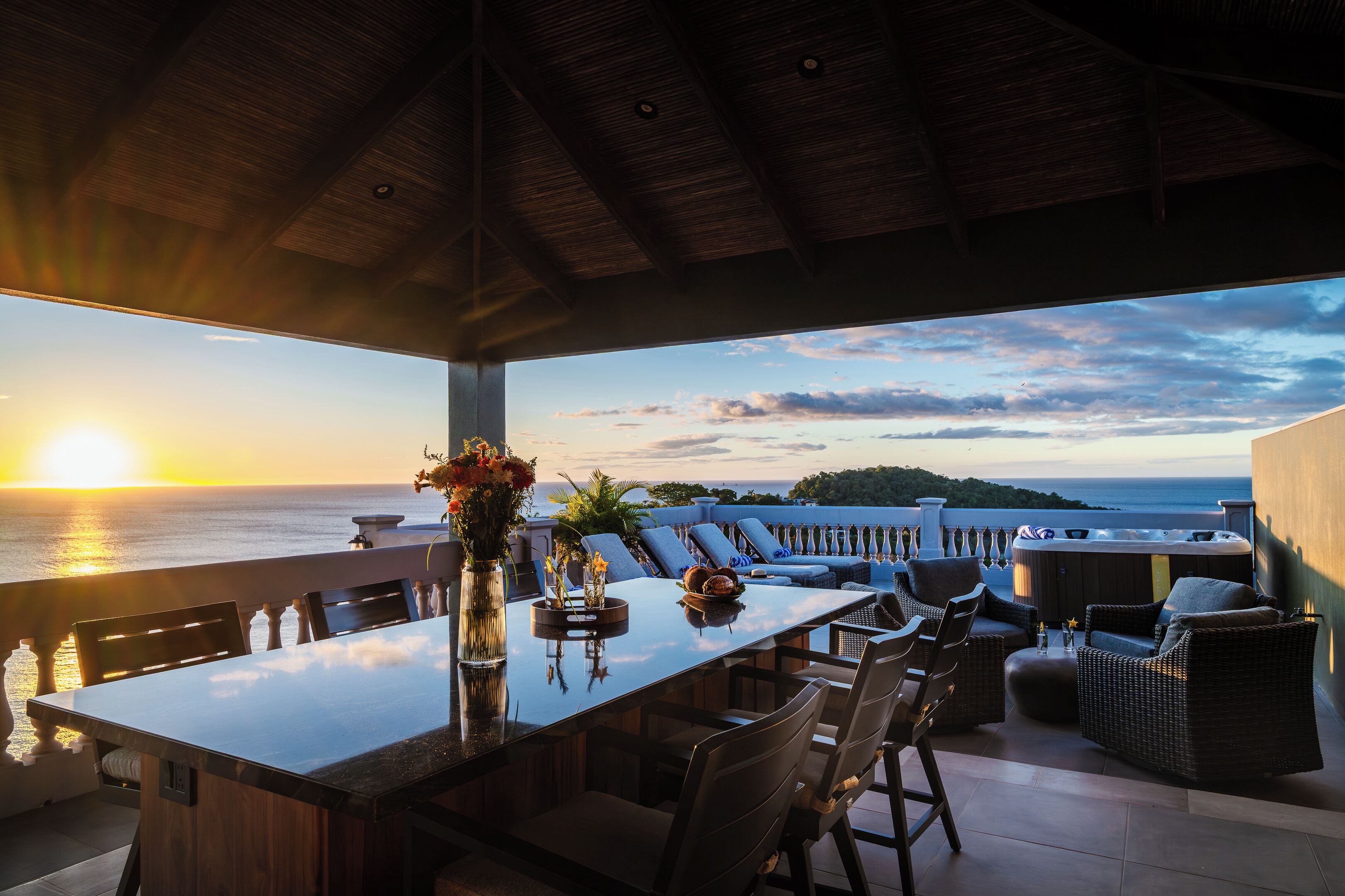 A terrace with a dining table, chairs, and a hot tub overlooking the sea at sunset.